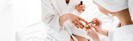 panoramic shot of women in bathrobes and jewelry sitting on bed and polishing nailsの写真素材
