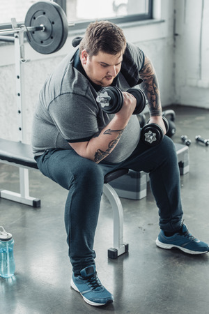 Overweight tattooed man sitting and exercising with dumbbells at sports centerの写真素材