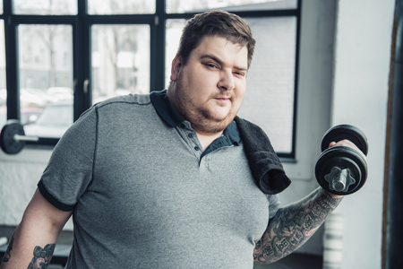 Overweight tattooed man with towel exercising with dumbbell and Looking At Camera at sports centerの写真素材
