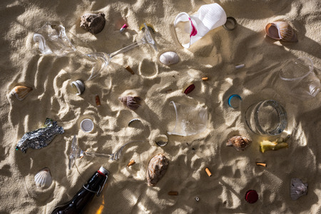 top view of seashells, glass bottle, scattered cigarette butts, broken glasses, apple core, plastic cups and candy wrapper on sand with shadowsの写真素材