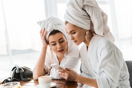 Stylish women in bathrobes and jewelry with towels on heads using smartphoneの写真素材