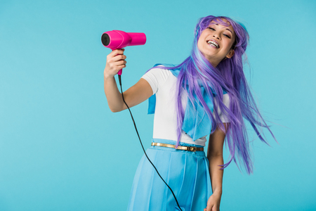 Smiling asian anime girl in wig using hairdryer isolated on blueの写真素材