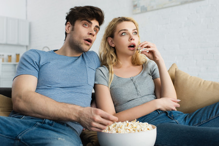 shocked man watching movie near blonde girl and bowl with popcornの写真素材
