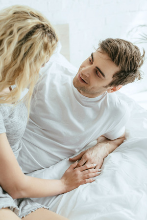 handsome and cheerful man looking at blonde girl lying on bedの写真素材