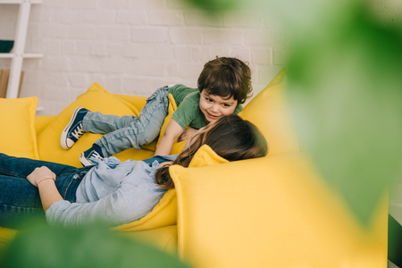 little boy playing with tired mother while she lying on yellow sofa in living roomの写真素材