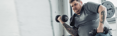 panoramic shot of Overweight tattooed man training with dumbbells at sports centerの写真素材