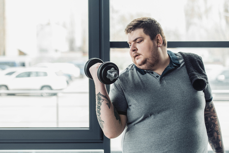 Overweight tattooed man with towel exercising with dumbbell at gym with copy spaceの写真素材