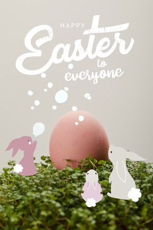 Pink painted chicken egg on green grass with happy Easter to everyone lettering and rabbits illustrationの写真素材