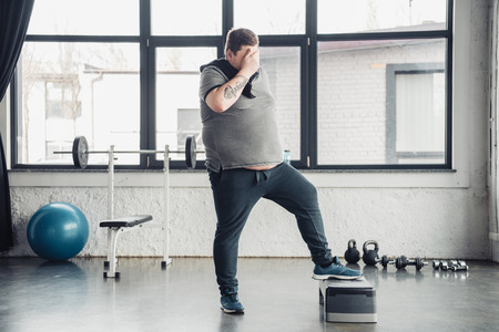 Overweight man with towel covering face with hand while training on step platform at sports centerの写真素材