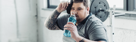 Panoramic shot of overweight tattooed man drinking water from sport bottle and wiping face with towel at gymの写真素材