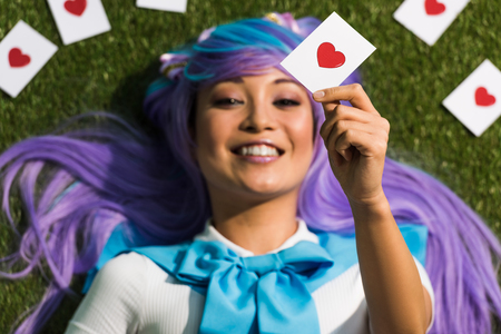 Selective focus of Asian anime girl in purple wig holding card and lying on grass