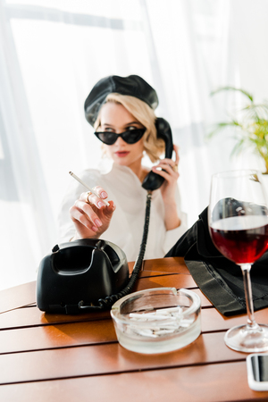 Selective focus of elegant blonde woman in black beret and sunglasses smoking and talking retro phone near glass with red wineの写真素材