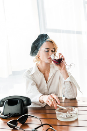 Elegant blonde woman in black beret and white shirt with closed eyes smoking cigarette and drinking red wineの写真素材