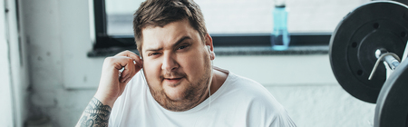 Panoramic shot of overweight man looking at camera while putting on earphones at gym
