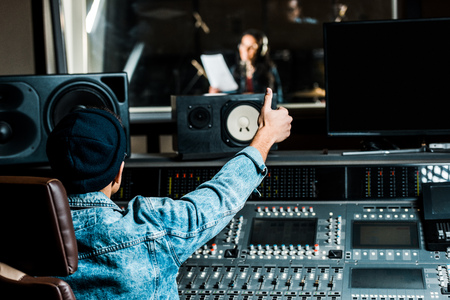 Selective focus of mixed race sound producer showing thumb up to woman singing in recording studioの写真素材