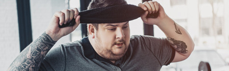 Panoramic shot of tired overweight tattooed man wiping face with towel at sports center