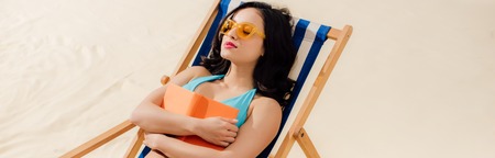 Panoramic shot of beautiful girl in bikini relaxing on deck chair with book on beach