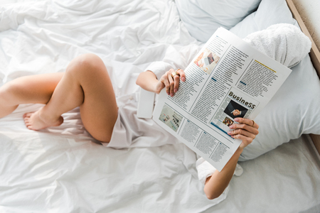 Top view of woman with towel on head and obscure face lying in white bed and reading business newspaperの写真素材