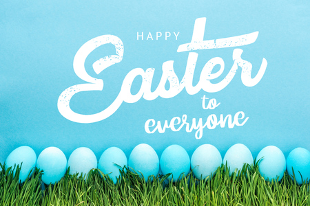 Blue painted chicken eggs on green grass with happy Easter to everyone lettering on blue background