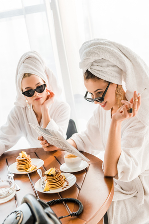 Stylish smiling woman in bathrobe, sunglasses and jewelry with towel on head smoking cigarette and reading newspaper with friend at morningの写真素材