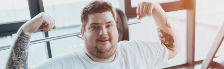 Panoramic shot of overweight tattooed man smiling and showing muscles at gym with sunlightの写真素材