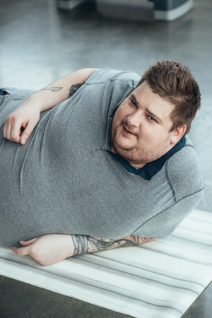 Overweight tattooed man in grey t-shirt looking at camera while lying on fitness mat at sports centerの写真素材