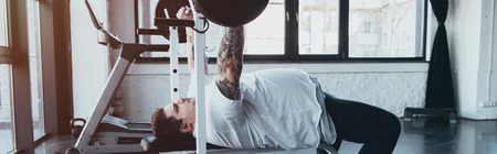 Panoramic shot of overweight tattooed man training with barbell at gym