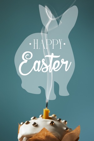 Selective focus of traditional Easter cake with fireless candle and smoke on blue background with happy Easter lettering and rabbit illustrationの写真素材
