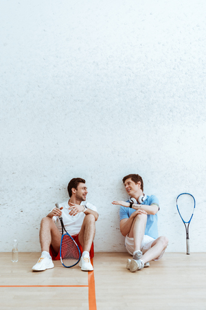 Smiling squash players sitting on floor and talking in four-walled courtの写真素材