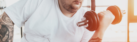 Panoramic shot of Overweight tattooed man in white t-shirt training with dumbbell at gymの写真素材