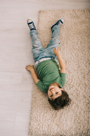 Overhead view of curious little boy in green t-shirt lying on carpetの写真素材