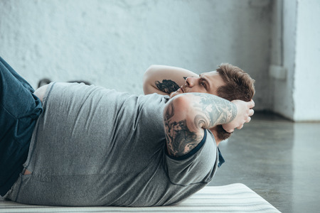 Overweight tattooed man lying on fitness mat and doing sit up exercise at sports centerの写真素材