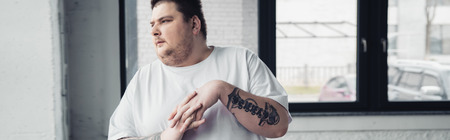 Panoramic shot of overweight tattooed man stretching fingers at gym