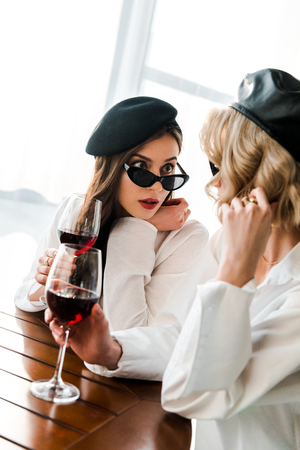 Elegant shocked brunette woman in black beret and sunglasses drinking red wine at wooden table with blonde friendの写真素材