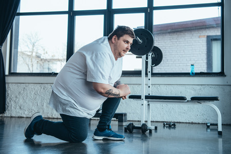 Overweight tattooed man in white t-shirt stretching at sports center