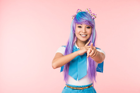 Smiling Asian anime girl in purple wig pointing with finger at camera isolated on pink background