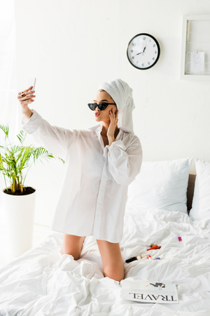 Stylish woman in shirt, sunglasses, jewelry and with towel on head taking selfie in bedの写真素材