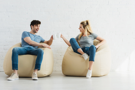 Attractive blonde girl and cheerful man holding cups while sitting on bean bag chairsの写真素材