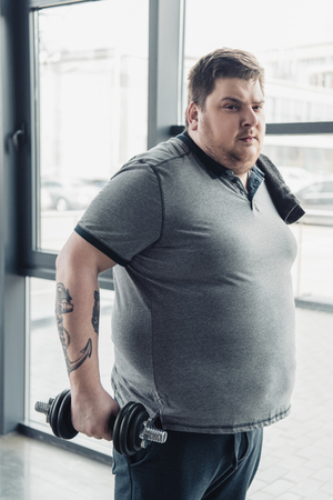 Overweight tattooed man in grey t-shirt with towel exercising with dumbbell at sports centerの写真素材