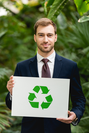 Handsome smiling businessman in suit holding card with green recycling sign in greenhouseの写真素材