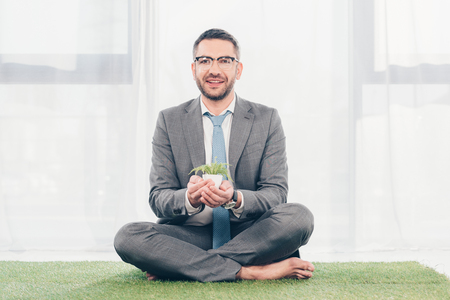 Handsome smiling businessman in suit sitting on grass mat and holding plant in flowerpotの写真素材