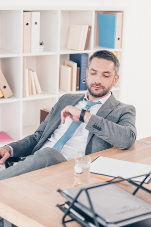Handsome businessman sitting at desk, looking at watch and checking time in officeの写真素材