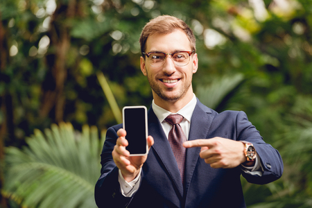 Smiling businessman in suit, tie and glasses pointing with finger at smartphone blank screen in greenhouseの写真素材