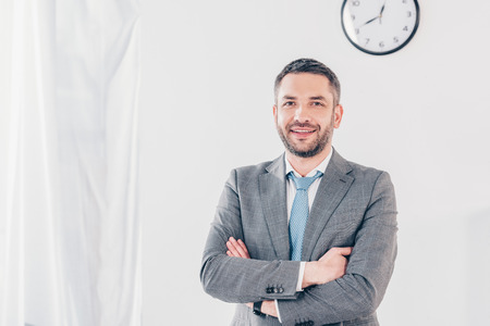 Handsome smiling businessman in suit with crossed arms looking at camera in office with copy spaceの写真素材