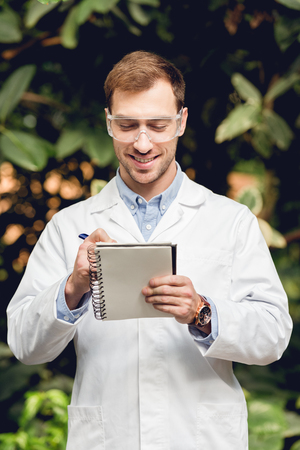 Smiling scientist in white coat and goggles making notes in green orangeryの写真素材