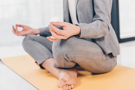 Partial view of businesswoman in suit sitting in Lotus Pose on fitness mat and meditating in officeの写真素材