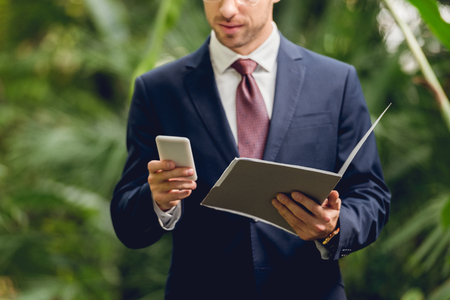 Cropped view of businessman in suit using smartphone and holding folder in greenhouseの写真素材