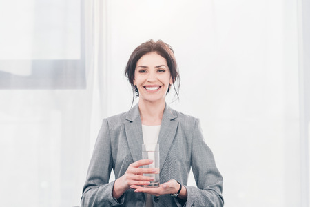 Beautiful smiling businesswoman in suit holding glass of water and looking at cameraの写真素材