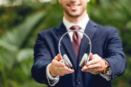 Partial view of smiling businessman in suit and tie holding wireless headphones in greenhouseの写真素材