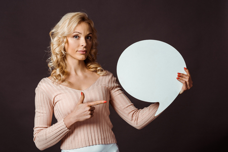 attractive blonde woman pointing with finger at speech bubble isolated on blackの写真素材